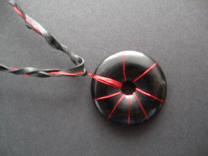 A round donut of obsidian stone wrapped in metallic red wire in a design similar to a firework on a braided cord of black cord and red wire.