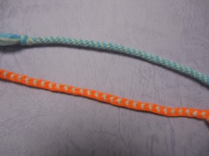 A braided cord of blue and  white yarn in stripes and a cord of orange with white hearts.