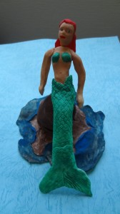 A mermaid with red hair sits on a brown rock over dark blue water on a light blue piece of paper.