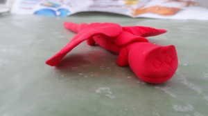 A small red clay dragon with diamond eyes, short stubby legs, and a long straight tail.