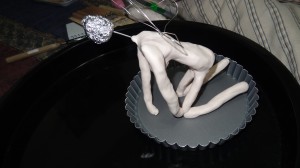 A human female figure kneeling with skin-colored clay from the neck down and wings coming from her back.  Her neck is a piece of wire and her head is a ball of aluminum foil.